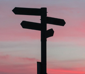 Schwarzes Wegweiser Schild mit Pfeilen und Sonnenuntergang im Hintergrund
