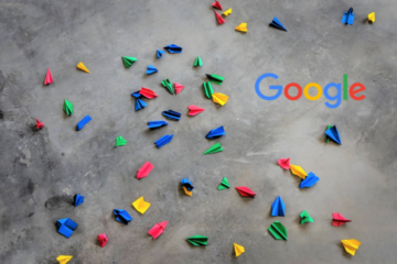 Kleine bunte Papierflieger und das Google Logo auf einer grauen Fläche