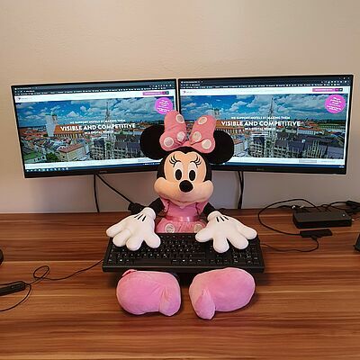 Minnie Maus als Plüschtier mit der Tastatur auf sich vor zwei Bildschirmen